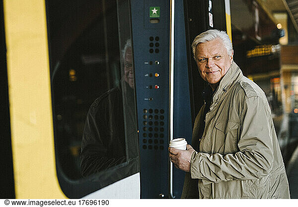 Side view of happy businessman holding disposable cup while boarding in train at railroad station