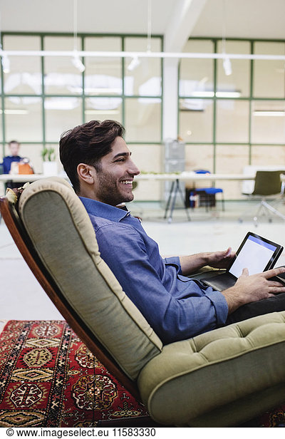 Side view of happy male blogger holding digital tablet while sitting on armchair in office