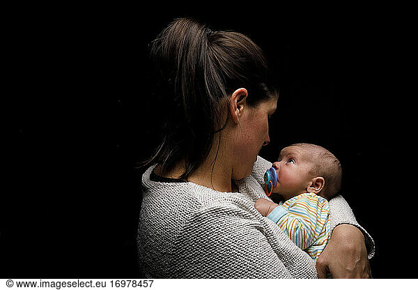 Side view of happy mom cuddling adorable newborn baby and enjoying moments of motherhood against black background