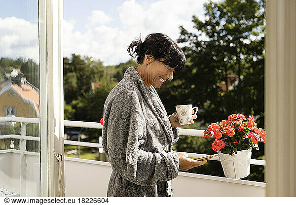 Side view of happy woman holding cup using smart phone while standing in balcony