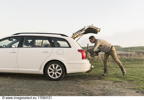 Side view of hiker searching in car trunk on field against clear sky