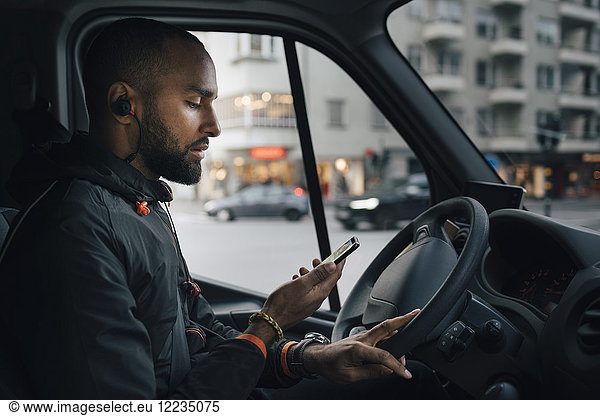Side view of male driver looking at mobile phone while sitting in delivery van at city