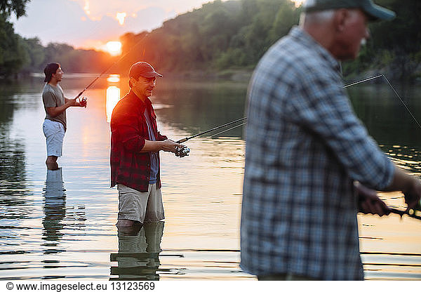 Side view of male friends fishing while standing in lake during sunset