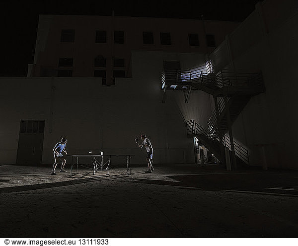 Side view of male friends playing table tennis against illuminated building