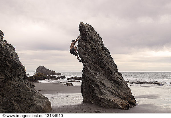 Side view of man climbing rock formation against cloudy sky