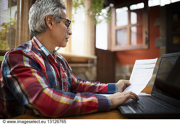 Side view of man looking and document and using laptop on table