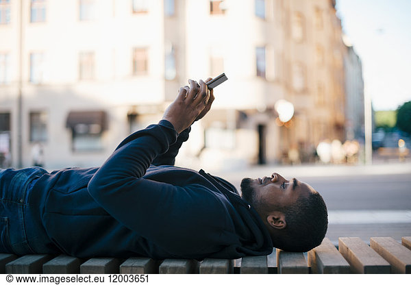 Side view of man lying on bench while using smart phone in city