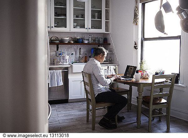 Side view of man using laptop computer while sitting at dining table in kitchen