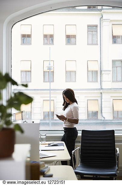 Side view of mature businesswoman using mobile phone while standing by window at office