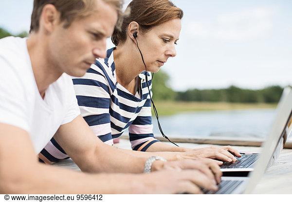 Side view of mature couple using laptops while lying on pier