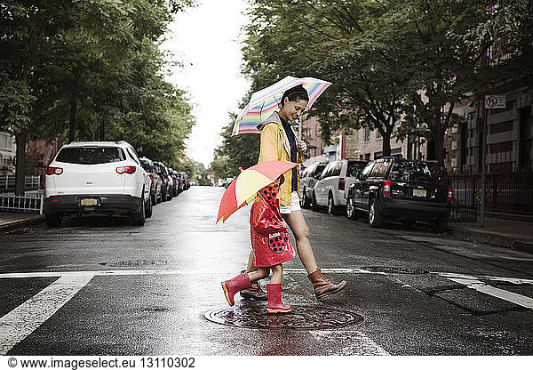 Side view of mother and daughter holding umbrellas while crossing road