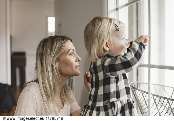 Side view of mother and daughter looking through window at home