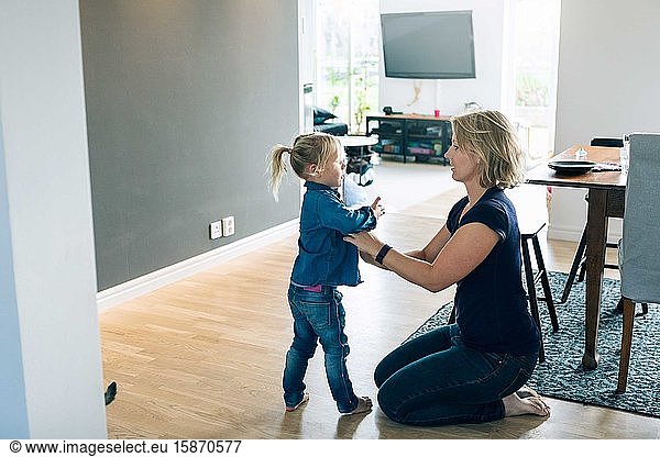 Side view of mother dressing daughter while kneeling on floor at home