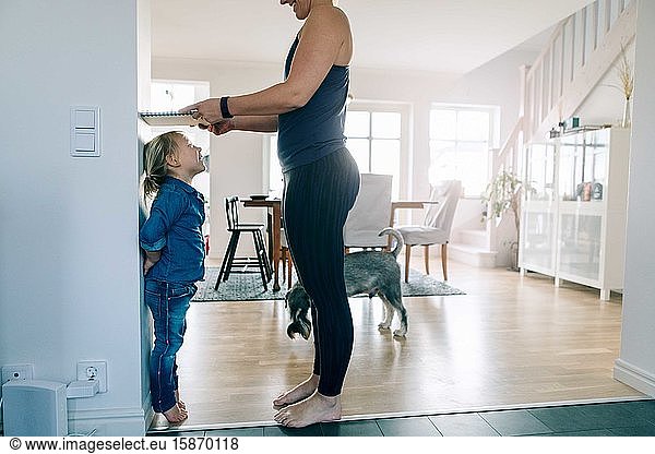 Side view of mother measuring daughter's height with book on wall at home