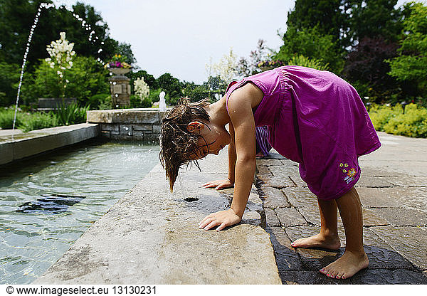 Side view of playful girl bending over fountain in park during sunny day