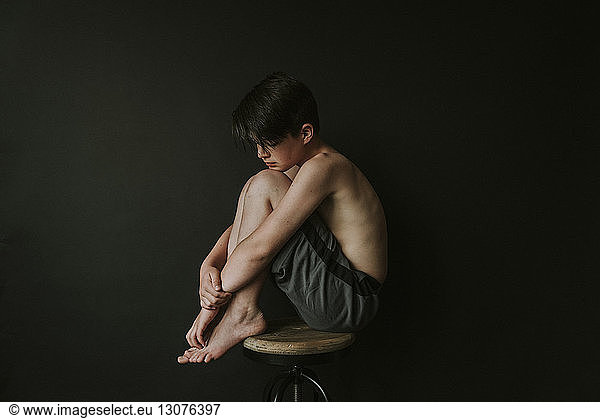 Side view of sad shirtless boy sitting on stool against black background