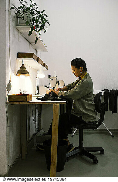 Side view of seamstress stitching clothes using sewing machine while sitting on chair at workshop
