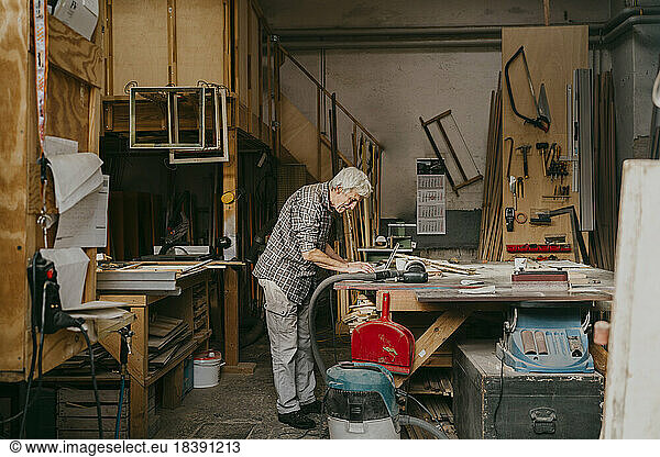 Side view of senior male carpenter working on laptop while standing at repair shop