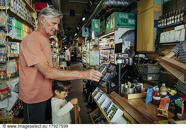 Side view of senior man doing online payment while standing with grandson at checkout counter in supermarket