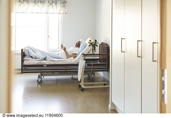 Side view of senior man using smart phone while reclining on bed in hospital ward
