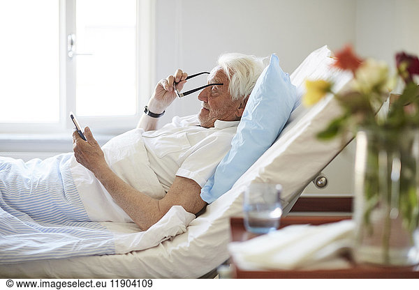 Side view of senior man wearing eyeglasses while using smart phone in hospital ward