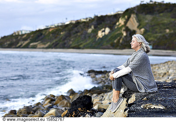 Side view of senior woman with eeditorial closed sitting on rock at Manhattan Beach during sunset