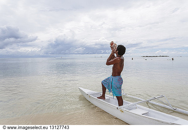 Side view of shirtless man blowing coach shell while standing in boat at beach against cloudy sky
