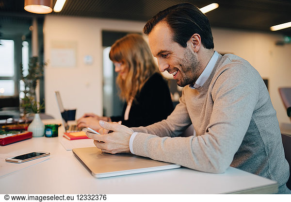 Side view of smiling businessman using smart phone while sitting by businesswoman at desk in creative office