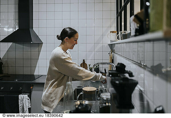 Side view of smiling businesswoman washing glass at sink in office kitchen