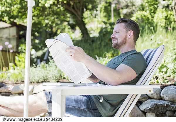 Side view of smiling man reading newspaper on lounge chair at yard
