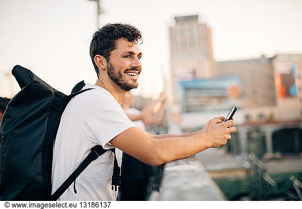 Side view of smiling young man looking away while holding mobile phone at bridge