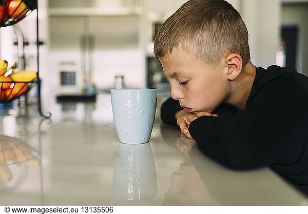 Side view of tired boy with tea cup resting head on kitchen counter at home