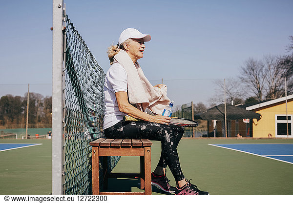 Side view of tired senior woman sitting on bench against sky at tennis court