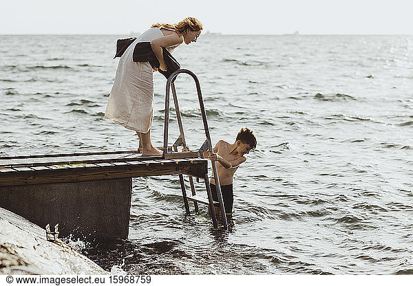 Side view of woman guiding while son moving down on ladder in sea