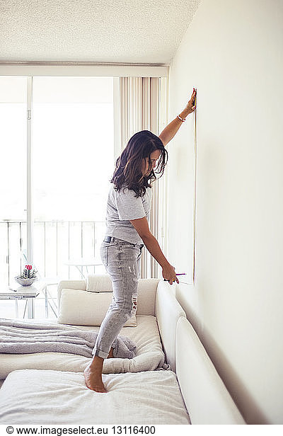 Side view of woman measuring wall while standing on sofa at home