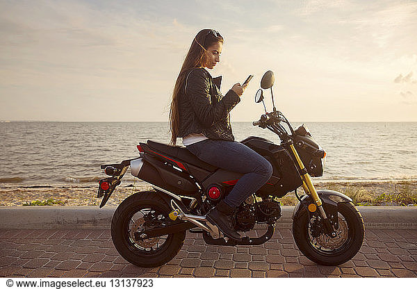 Side view of woman sitting on motorcycle and using smart phone against sea