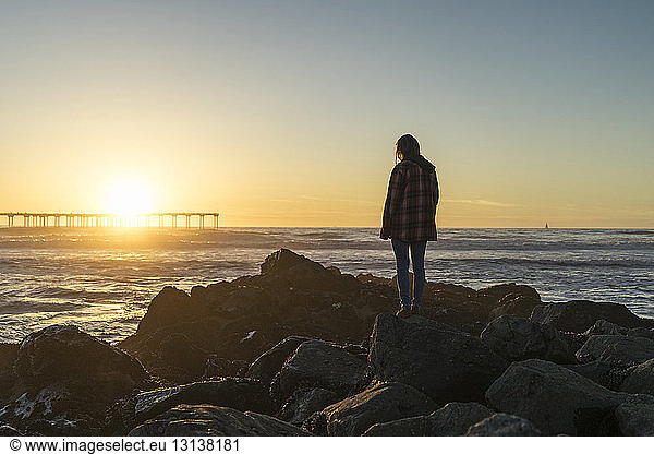 Side view of woman standing on rocky shore against sky during sunset