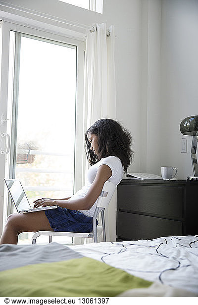Side view of woman using laptop while sitting on chair by window at home