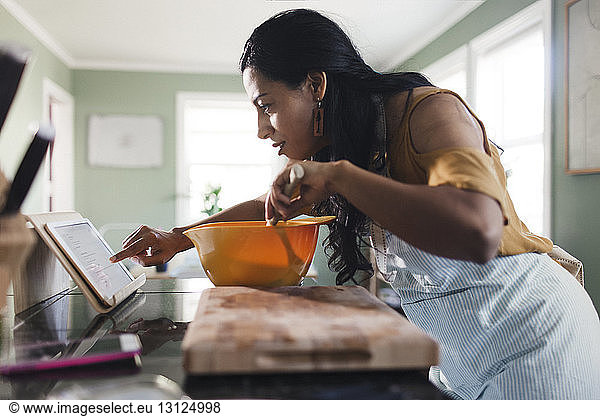 Side view of woman using tablet computer for recipe while preparing food in kitchen