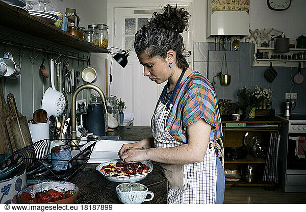 Side view of woman wearing apron garnishing pie while standing in kitchen