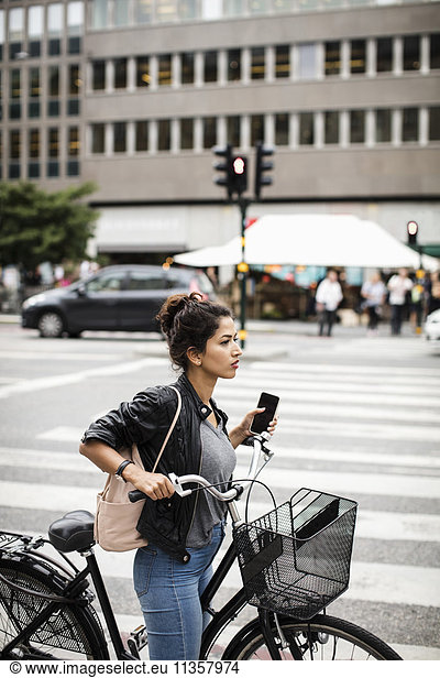 Side view of woman with bicycle holding phone while standing on zebra crossing in city