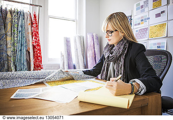 Side view of young female fashion designer checking fabric on desk at workshop