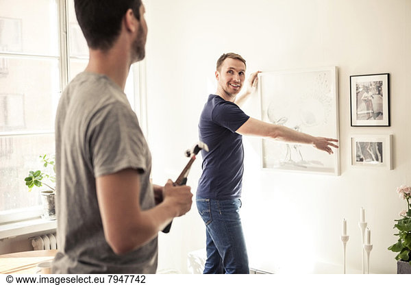 Side view of young gay man looking at partner while hanging frame on wall in home
