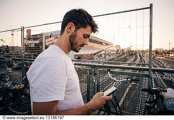 Side view of young man using mobile phone while standing on bridge over railroad tracks in city
