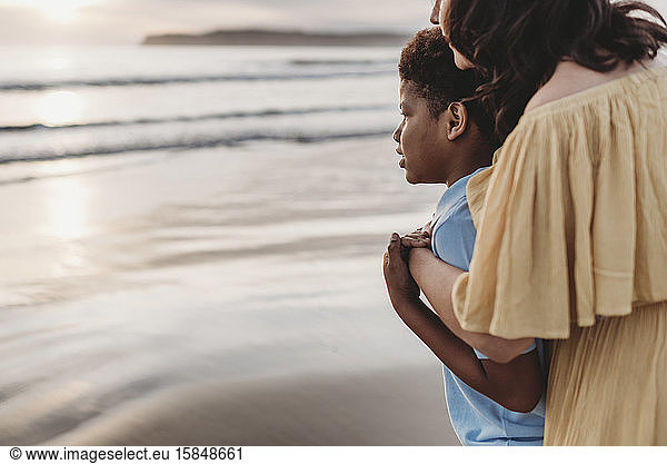 Side view of young mom embracing son at seashore during sunset