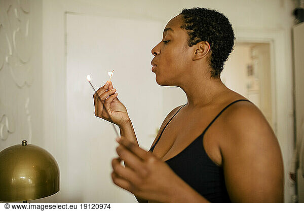 Side view of young woman blowing matchstick while holding incense at home