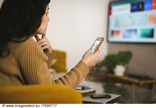 Side view of young woman controlling TV through mobile app at home