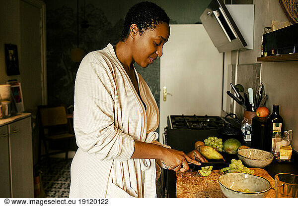 Side view of young woman cutting kiwi at kitchen counter