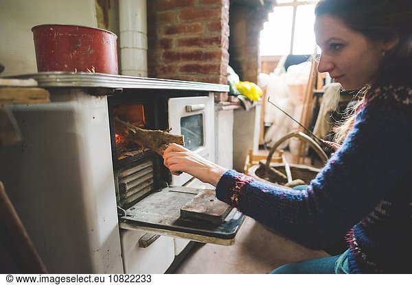Side view of young woman putting firewood into wood burner