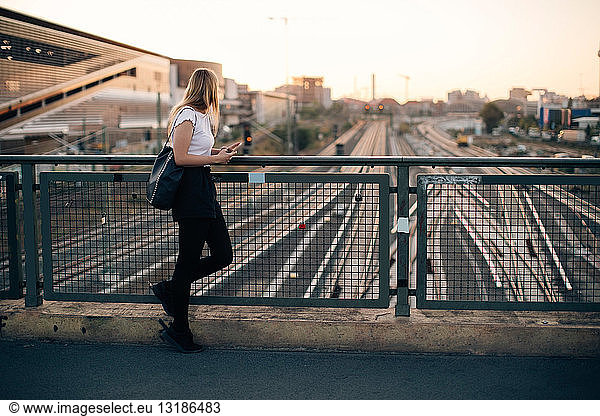 Side view of young woman standing on bridge over railroad tracks in city during sunset